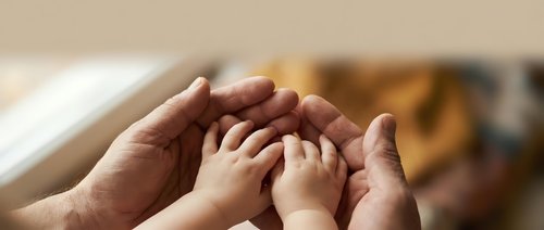 Adult and child hands forming protective circle together showing family care, support and unity in warm indoor setting.