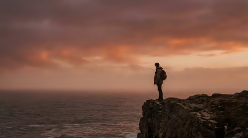 Silhouetted male hiker standing on rocky cliff overlooking vast ocean at sunset, wearing backpack. Dramatic golden and orange sky with clouds. Concept for adventure, travel, exploration, and outdoor lifestyle.
