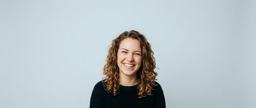 Young cheerful woman with curly brown hair laughing joyfully in black sweater against light gray background for lifestyle and portrait projects.