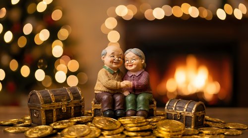 Elderly couple figurines sitting together with treasure chests and gold coins by warm fireplace with Christmas tree bokeh lights creating cozy holiday atmosphere.