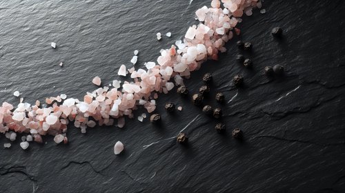 Pink Himalayan salt crystals scattered diagonally across dark slate stone background, creating dramatic contrast and texture for culinary and wellness concepts.