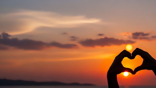 Hands forming heart shape silhouette against golden sunset sky over water. Romantic love symbol gesture at dusk with warm orange and pink clouds.