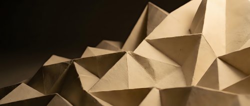 Abstract geometric paper origami sculpture with angular triangular facets creating dimensional shadows and depth on neutral background.
