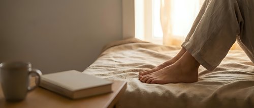 Woman relaxing on bed with book and coffee cup in warm morning sunlight streaming through window creating cozy bedroom atmosphere for rest and leisure.