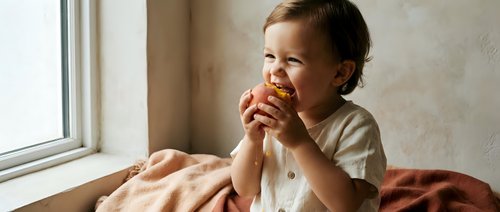 Young child, approximately 2-3 years old, Caucasian, sitting by window eating apple, wearing light colored shirt, natural daylight, indoor domestic setting.