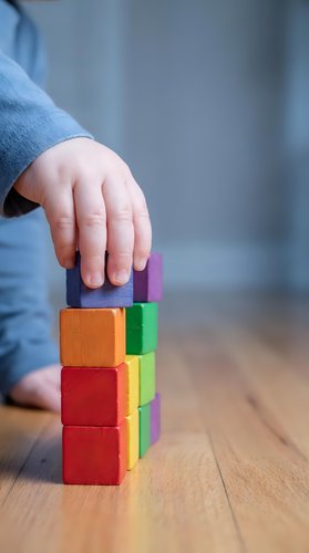 Child hand placing colorful wooden blocks in tower formation on wooden floor demonstrating early learning development and fine motor skills practice.