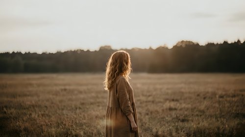 Young woman with long wavy hair standing in golden field during sunset, peaceful countryside moment with warm natural lighting and serene atmosphere.