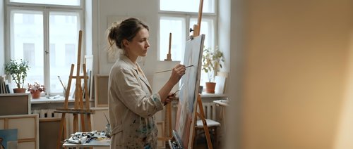 Young Caucasian woman artist painting on canvas in bright art studio workspace with easel and natural window light for creative projects.
