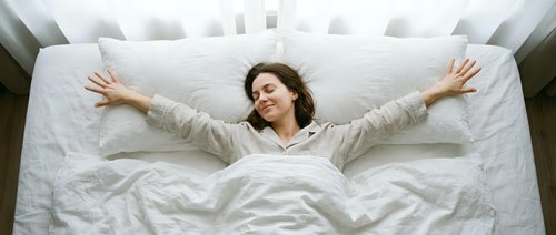 Woman with curly dark hair lying on white bedding with arms stretched out, expressing joy and relaxation. Overhead view for wellness and lifestyle content.