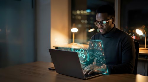 African American businessman working late at night on laptop with holographic digital interface technology in modern office environment.