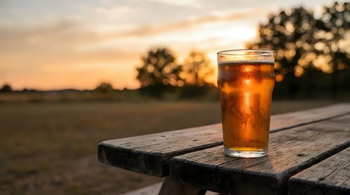 Cold refreshing beer glass on wooden table during golden hour sunset with countryside landscape background for relaxation and leisure concepts.