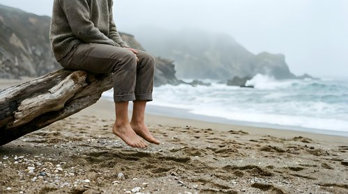 Person sitting on driftwood log at misty coastal beach with waves, barefoot relaxation and contemplation by ocean shore.