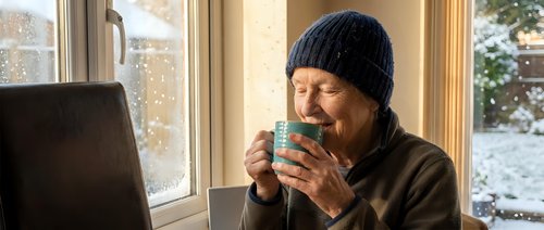 Mature Asian woman in knit beanie enjoying hot beverage by window on snowy winter day, cozy indoor lifestyle moment with laptop for remote work or relaxation.