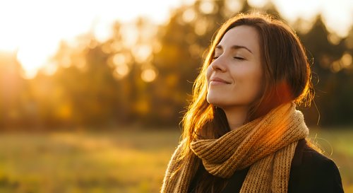 Young woman with closed eyes enjoying warm sunlight in autumn park wearing cozy knit scarf during golden hour sunset creating peaceful mindful moment outdoors.