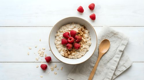 Creamy oatmeal porridge topped with fresh raspberries and granola in a ceramic bowl, wooden spoon on linen napkin, white wood background.