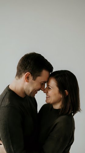 Happy young couple embracing intimately with foreheads touching, smiling warmly in cozy sweaters against neutral background for relationship content.