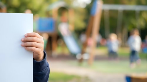 Child's hand holding blank white card with blurred playground background, suitable for advertising, marketing campaigns, educational materials, and promotional content.
