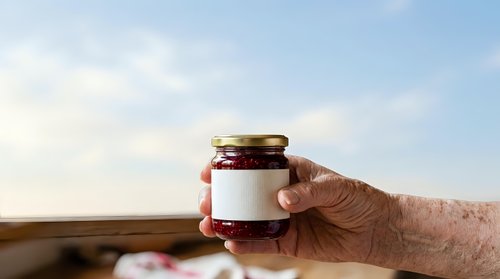 Elderly person with fair skin holding a glass jar with blank white label and dark red jam or preserve against rural landscape background for product packaging and branding.