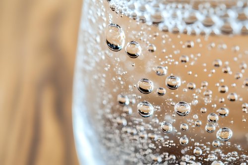 Macro shot of sparkling water bubbles in glass against wooden background, showing transparent spherical formations and natural refraction patterns.