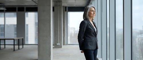 Mature woman in business attire standing by large windows in modern office space, looking out at cityscape. Corporate professional environment.
