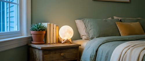 Cozy bedroom interior with warm lighting from bedside lamp, sage green pillows, books and plant on wooden nightstand creating peaceful atmosphere.