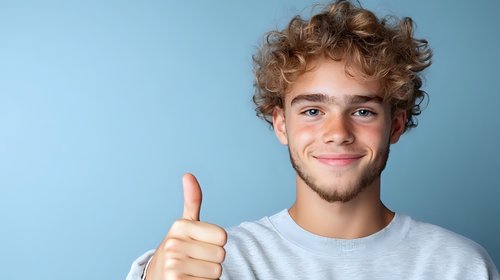 Young Caucasian man with curly blonde hair and friendly smile showing thumbs up gesture against blue background, wearing casual white t-shirt.