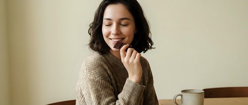 Young Asian woman in cozy beige sweater enjoying peaceful moment with eyes closed, hand near chin in contemplative pose. Coffee cup on wooden table creates warm atmosphere.