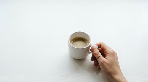 Hand holding white ceramic mug with hot coffee on white background, overhead view for beverage, lifestyle and morning routine concepts.