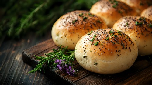 Fresh baked bread rolls with poppy seeds and herbs on rustic wooden cutting board, garnished with dill and purple flowers in dark moody setting.