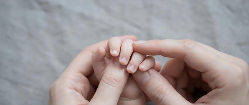 Adult hands gently holding tiny newborn baby fingers in tender moment of connection and care on soft gray background.