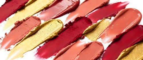 Colorful lipstick smears in red, pink, coral and gold tones creating artistic makeup texture pattern on white background for beauty and cosmetics design.