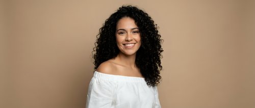 Young mixed-race woman with curly hair smiling warmly in white off-shoulder top against neutral beige background for lifestyle and portrait photography.