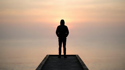 Silhouetted male figure standing on wooden pier at sunrise or sunset, gazing toward calm water horizon. Contemplative scene suitable for motivational, inspirational, and lifestyle content.