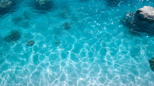Crystal clear turquoise ocean water surface with sunlight reflections creating natural ripple patterns, rocks visible beneath transparent azure waters.