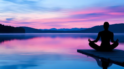 Silhouette of person meditating on wooden pier at sunset, purple and pink sky reflects in calm lake water surrounded by mountain landscape.