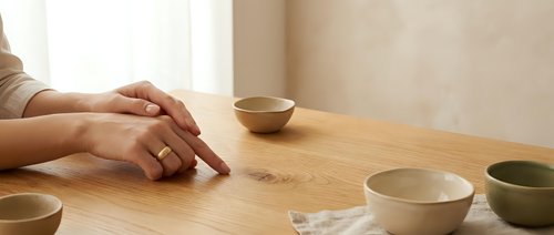 Woman hands resting on wooden table with ceramic bowls in minimalist kitchen interior. Natural lighting creates calm atmosphere for mindful living.