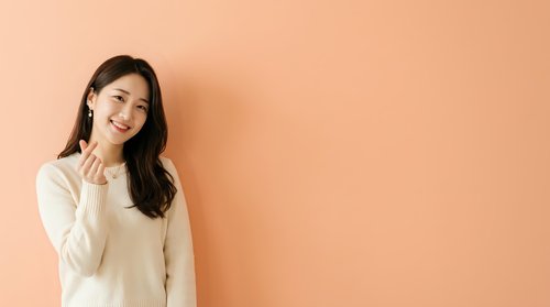 Young Asian woman with long dark hair wearing a cream-colored sweater, smiling and waving against a warm peach background. Suitable for lifestyle, portrait, and commercial advertising projects.