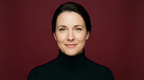 Professional Caucasian businesswoman in black turtleneck smiling confidently against burgundy background for corporate headshot and marketing materials.
