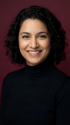 Professional Middle Eastern woman with curly hair smiling confidently in black turtleneck against burgundy background for business and corporate use.