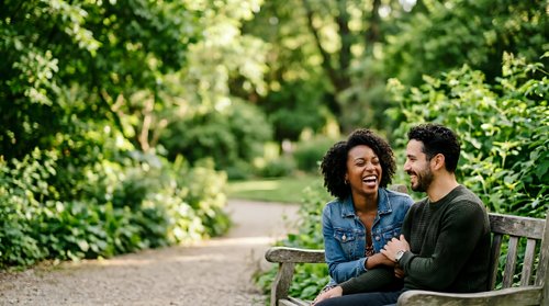 Young couple sitting on wooden bench in lush green park, enjoying conversation and connection outdoors during daytime. Woman with curly dark hair wearing denim jacket, man with beard in olive shirt.