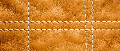 Brown leather texture with white decorative stitching in grid pattern. Natural grain surface with contrasting thread details for fashion and craft projects.
