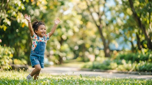 Young child in denim overalls running outdoors with arms raised in joy on sunny park path surrounded by green trees and natural foliage.