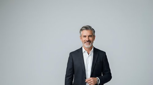Middle-aged man with gray hair and mustache wearing dark blazer and white shirt, standing against neutral background for corporate portraits and business headshots.