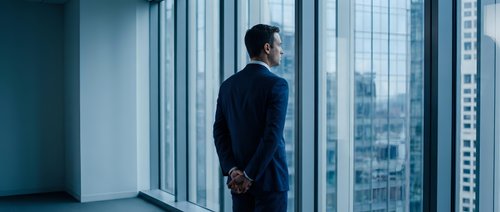 Professional businessman in dark suit standing by floor-to-ceiling windows overlooking urban cityscape, contemplating future business decisions and opportunities.