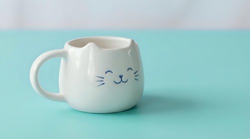 White ceramic cat-shaped mug with cute face and ears on mint green background. Adorable kitty coffee cup for pet lovers and kawaii style enthusiasts.