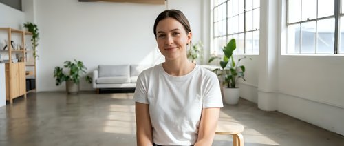 Smiling young Caucasian woman in white t-shirt sitting in bright modern apartment with large windows and plants, natural lighting for lifestyle content.