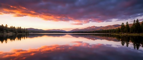 Serene lake at sunrise with vibrant orange and purple sky reflecting on calm water, surrounded by evergreen forest and distant mountains, peaceful nature landscape.