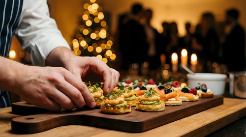 Chef preparing gourmet holiday appetizers on wooden board with Christmas tree lights and dinner party guests in warm restaurant setting.