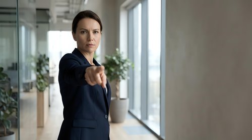 Professional businesswoman pointing finger at camera in modern office environment with confident expression and formal attire.
