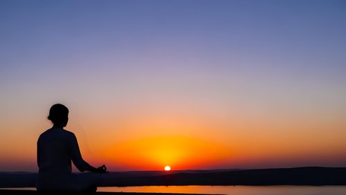 Silhouette of person meditating in lotus position during golden hour sunset over calm water with colorful sky gradient for wellness and mindfulness concepts.